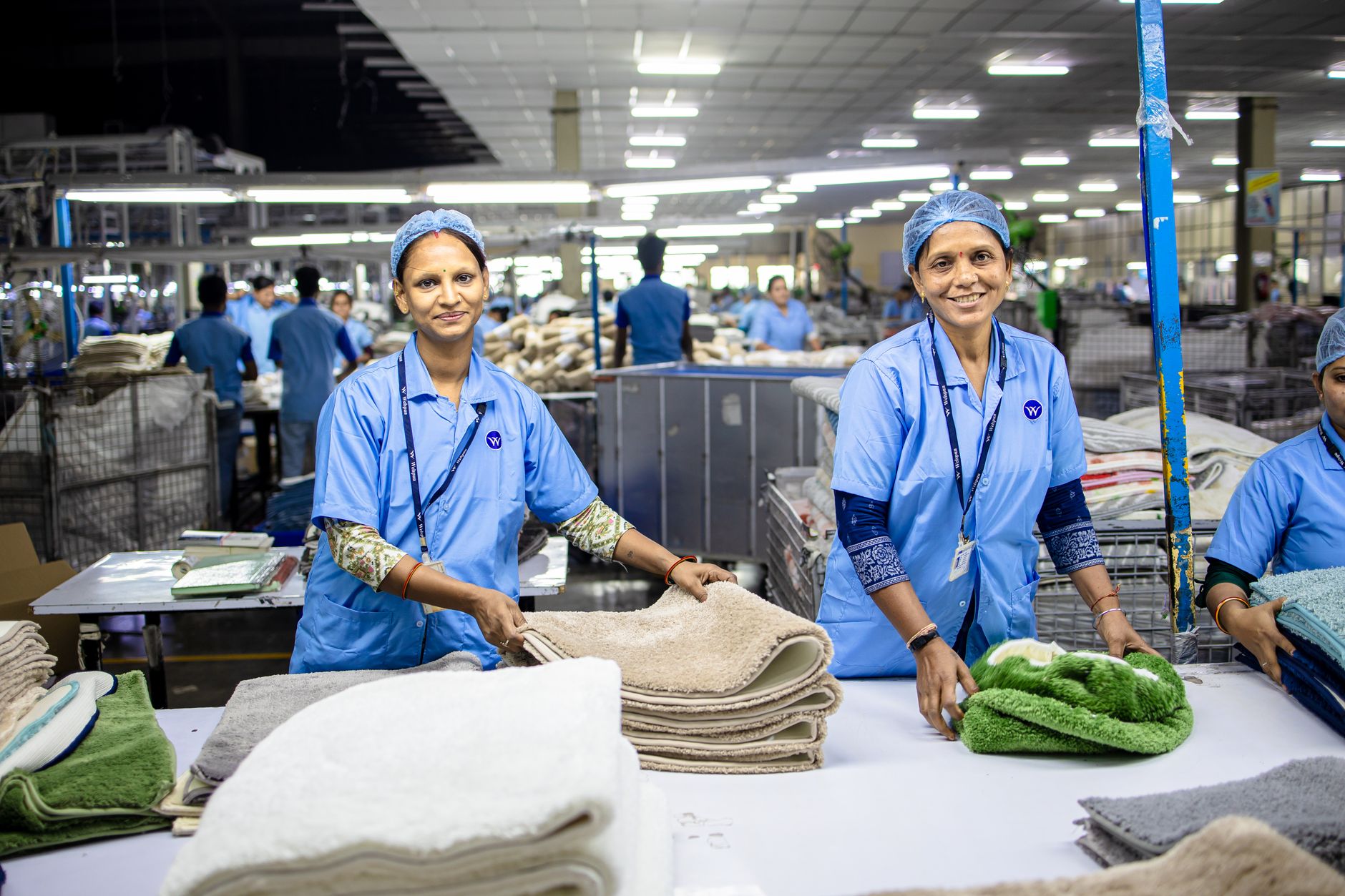 Deux employées en blouse bleue et charlotte plient des serviettes éponge sur une table dans un grand atelier textile.