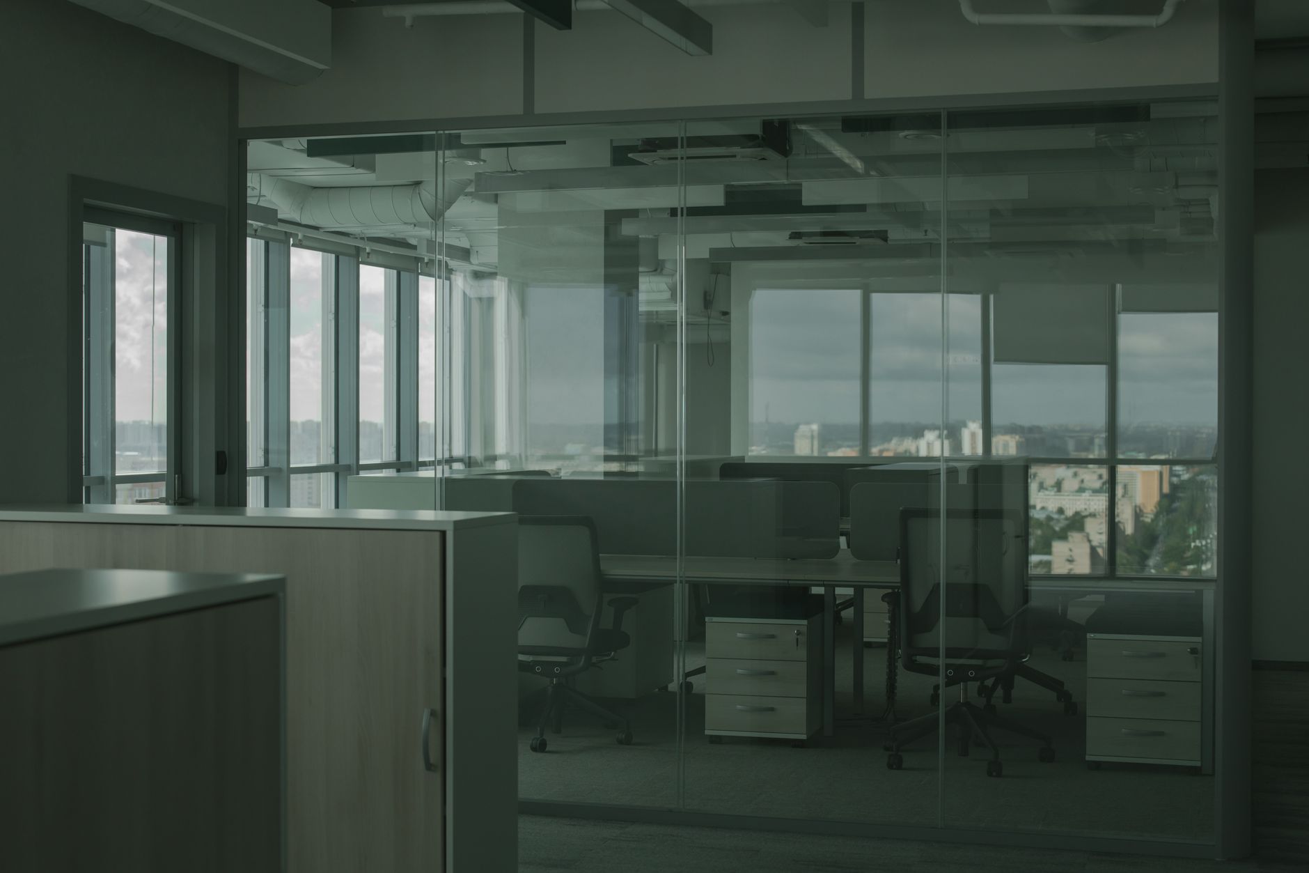 Open space de bureaux vide derrière une cloison vitrée, avec chaises, caissons à tiroirs et grandes fenêtres donnant sur la ville.
